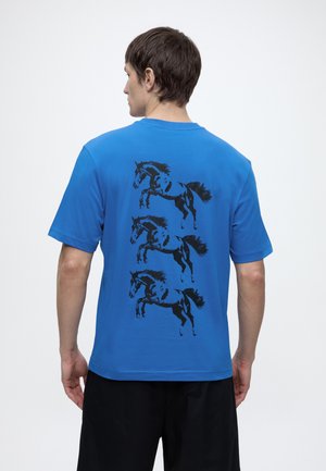 Uomo che indossa una maglietta blu con tre grafiche nere di cavalli in fila verticale sulla schiena, abbinata a pantaloncini neri.