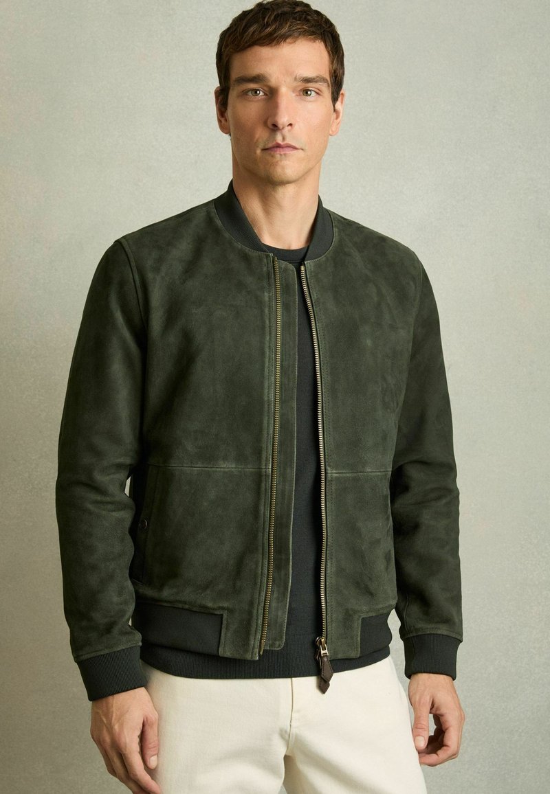 Jachetă bomber din suede verde închis, cu fermoar frontal, manșete și tiv din tricot. Prezintă o textură moale și elemente de design minime.