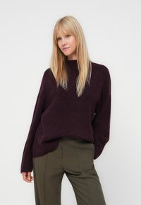 Burgundy neulepaita rentoon leikkaukseen, korkealla kauluksella ja pitkillä hihoilla, yhdistettynä oliivinvihreisiin räätälöityihin housuihin. Pehmeä materiaali ja minimalistinen muotoilu.