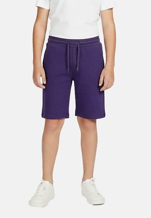 Kind draagt paarse shorts met trekkoord, witte sneakers en een wit T-shirt, staande tegen een effen achtergrond.