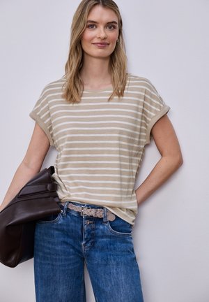 Donna che indossa una maglietta a maniche corte a righe beige e bianche e jeans blu, che tiene in mano una giacca marrone scuro piegata davanti a uno sfondo monocromatico.