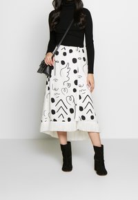 Kvinna i svart polotröja, vit midikjol med svarta abstrakta ansikten och prickar, svarta klackstövlar och liten svart axelväska.