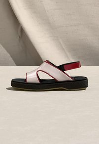 Ανοικτό σανδάλι με επάνω μέρος από λευκό δέρμα, κόκκινες λεπτομέρειες, μαύρη επενδεδυμένη σόλα και πίσω λουράκι, τοποθετημένο σε ανοιχτόχρωμη επιφάνεια με ουδέτερο φόντο.