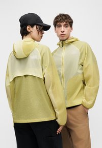 Deux personnes portent des vestes légères à capuche jaunes, l'une faisant face avec les mains dans les poches, l'autre montrant le dos avec une casquette noire.