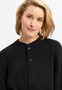 Schwarze Strickjacke mit rundem Ausschnitt, versehen mit einem Knopfverschluss und strukturiertem Stoff. Das Design hat eine lockere Passform und ein weiches Gefühl.