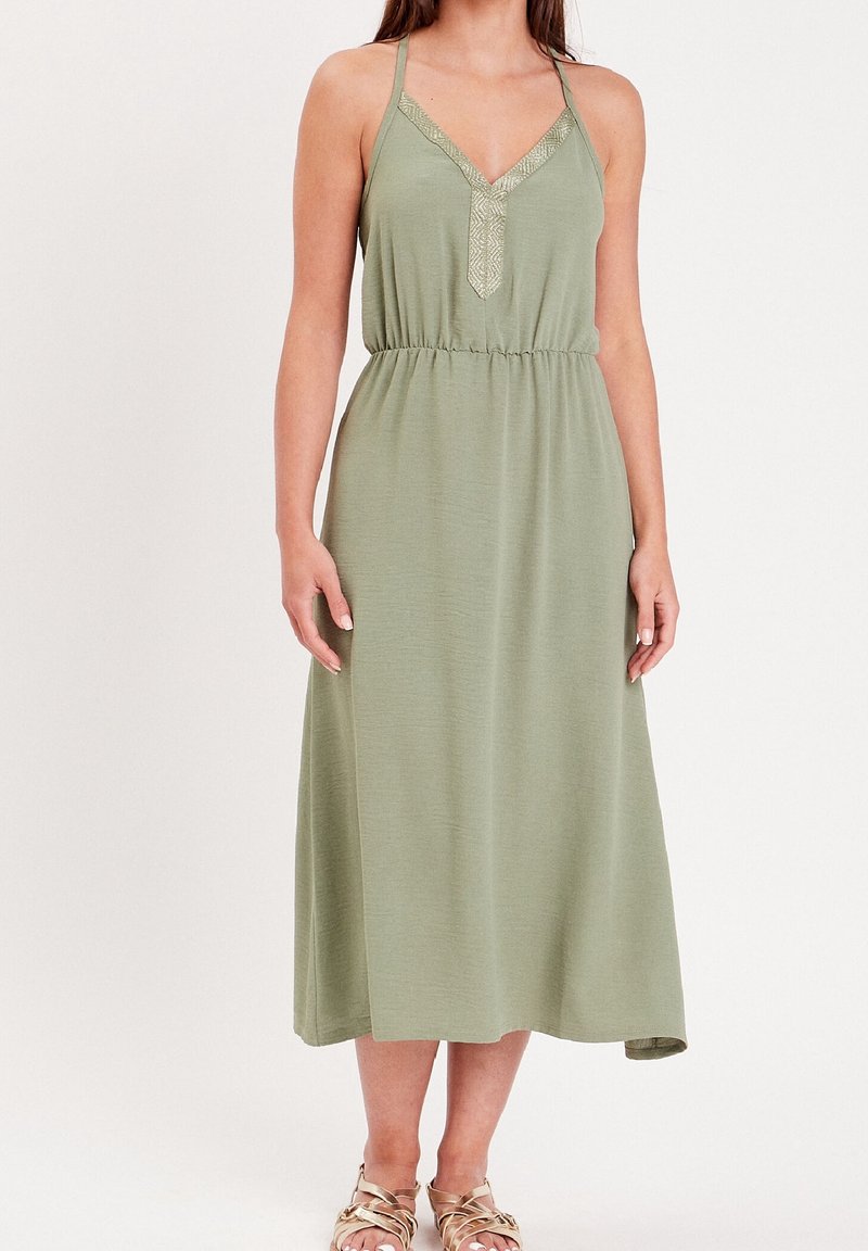 Femme portant une robe midi sans manches vert clair avec bordure en dentelle au col en V et des sandales dorées à brides, se tenant devant un fond uni.