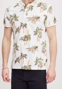 Witte polo met een groen, bruin en oranje palmboomprint. Heeft een kraag en korte mouwen. Etiket in de linkerbovenhoek.