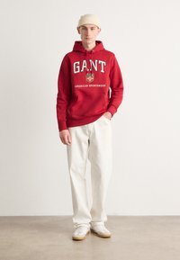 Sudadera de algodón roja con estampado blanco "GANT" y "AMERICAN SPORTSWEAR", combinada con pantalones baggy beige claros y zapatillas blancas.