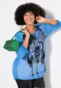 Chemise à manches longues bleue avec impression graphique et accents noirs, assortie à un sac à main texturé vert. Le mannequin pose sur un fond blanc.