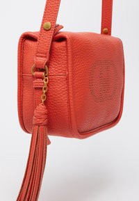 Červená textúrovaná kožená crossbody taška obdĺžnikového tvaru so zlatými kovovými detailami a aplikáciou strapca. Na prednej strane má perforovaný detail loga.