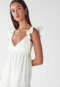 Valkoinen mekko, jossa on röyhelöhihoja, syvä V-kaula-aukko ja laskostettu vyötärö. Kevyt kangas, virtaava hame, sileä tekstuuri.