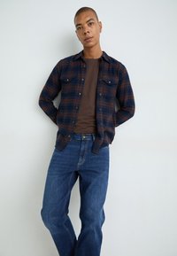 Cămașă din flanel cu carouri navy și rust, cu două buzunare pe piept, purtată peste un tricou bej cu guler rotund, asortată cu blugi drepti de culoare albastră.