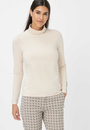 Frau mit langen dunklen Haaren trägt einen beigen Rollkragenpullover und gemusterte Hose, steht mit einer Hand in der Tasche vor einem schlichten weißen Hintergrund.