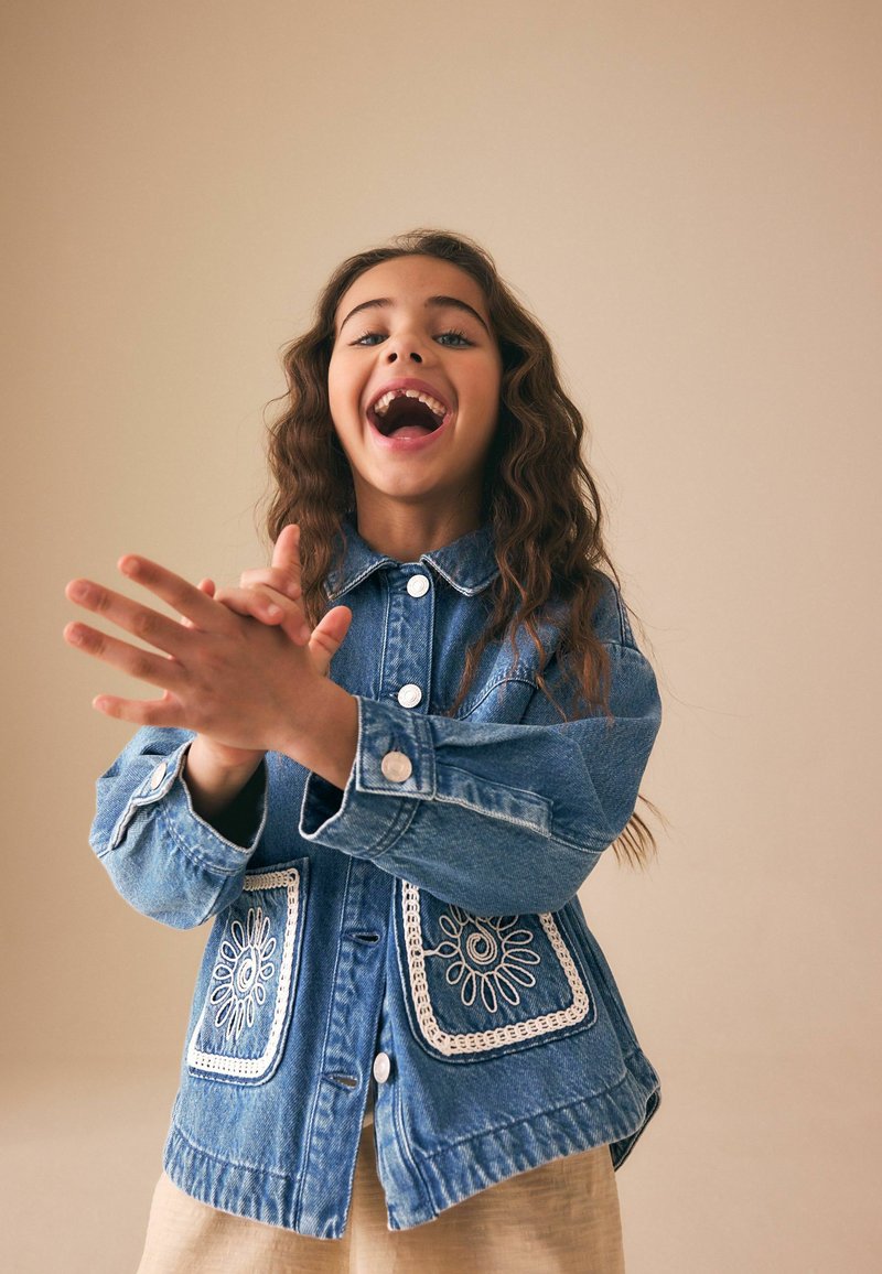 Young girl with long curly hair smiling widely and clapping hands, wearing a blue denim jacket with decorative white pockets.