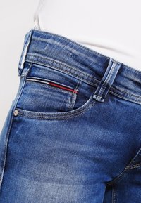 Vaqueros de mezclilla azul con una cintura alta. Las características incluyen un detalle de rayas rojas y negras en el bolsillo delantero y costuras en contraste.