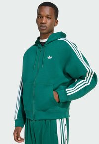 Uomo che indossa una felpa con zip verde Adidas e pantaloni da jogging coordinati con strisce bianche, in piedi con una mano in tasca su uno sfondo semplice.