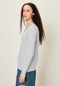 Pelēks krekliņš ar garām piedurknēm un V veida izgriezumu, ar mīkstu tekstūru un piegulošām piedurknēm, kombinēts ar zilām augsta vidukļa džinsu biksēm ar izšūtu plāksteri.