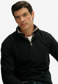 Młody mężczyzna z ciemnymi włosami, ubrany w czarny sweter z kołnierzykiem na zamek, nałożony na koszulę w kratę i białą koszulkę pod spód, siedzący na tle jednolitym.