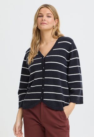 Kvinde iført en marineblå og hvidstribet cardigan med knapper og trekvartærmer samt bordeaux bukser, står med en hånd i lommen.