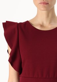Burgundin mekko, jossa on rypytetyt hihat, pyöreä kaula-aukko ja sileä, tekstiililtään vaihteleva kangas. Keskitytään ylävartalon yksityiskohtiin.