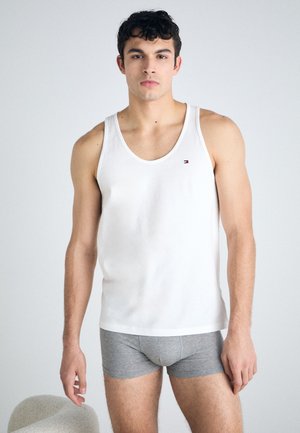 Débardeur blanc sans manches avec un col rond et petit logo sur la poitrine. Porté avec un boxer gris. Le modèle se tient devant un fond clair.
