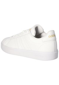 Biele športové tenisky s textúrovaným syntetickým zvrškom, zaoblenou špičkou, tromi bočnými pruhmi a zlatými logovými akcentmi. Rovná biela podrážka.