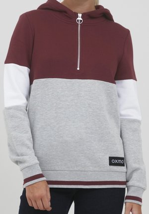 Osoba nosząca bluzę z kapturem w kolorowe panele w kolorach bordowym, białym i szarym, z półzamkiem z przodu i etykietą "oxmo" w dolnym prawym rogu.