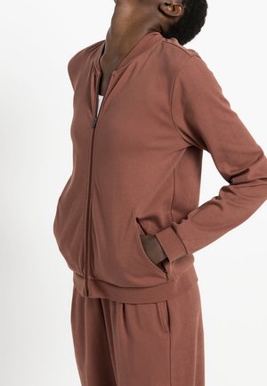 Persona con chaqueta marrón con cremallera y pantalones a juego, con una mano en el bolsillo de la chaqueta y la cabeza inclinada hacia atrás.