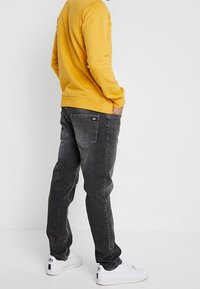 Henkilö, jolla on päällään sinapinkeltainen villapaita, mustat haalistuneet farkut ja valkoiset lenkkarit, seisoo kädet taskuissa yksinkertaista valkoista taustaa vasten.