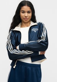 Jeune femme aux longs cheveux foncés portant une veste Adidas Originals bleu marine et un pantalon assorti, les bras croisés, debout devant un fond uni.