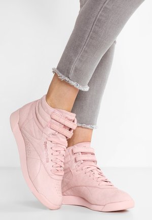 Osoba nosząca jasnoróżowe wysokie trampki Reebok z podwójnymi paskami oraz jasnoszare jeansy z postrzępionym mankietem na białym tle.