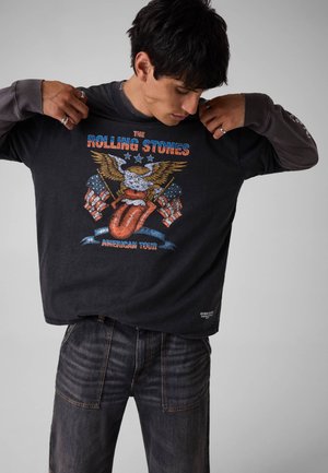 Junger Mann richtet ein schwarzes Rolling Stones American Tour Shirt mit einem Adler- und amerikanischen Flaggen-Design.