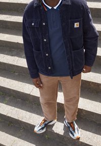 Giacca di velluto a coste blu navy con tasche frontali sopra una polo blu, abbinata a pantaloni a strisce marroni e scarpe sportive con dettagli arancioni.