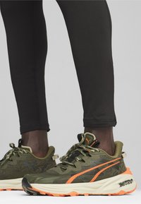 Zielono-czarne buty sportowe z pomarańczowymi akcentami, teksturowana cholewka, gruba podeszwa, wzorzyste detale i odblaskowe sznurowadła.