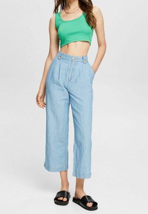 Frau trägt grünes, bauchfreies Top, hellblaue, weit geschnittene Hose mit hoher Taille und schwarze Slide-Sandalen vor einfachem Hintergrund.