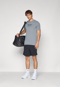 Grå Nike Pro T-skjorte med svart logo, svarte shorts, hvite sneakers og hvite sokker. Modellen holder en svart treningsbag.