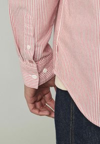 Chemise à rayures rouges et blanches, à manches longues, avec des poignets à un bouton et un ourlet légèrement arrondi, assortie à un jean foncé.