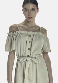 Vestido beige de un hombro con mangas con volantes, con botones en la parte delantera y una cintura atada. Tejido suave con una textura lisa.