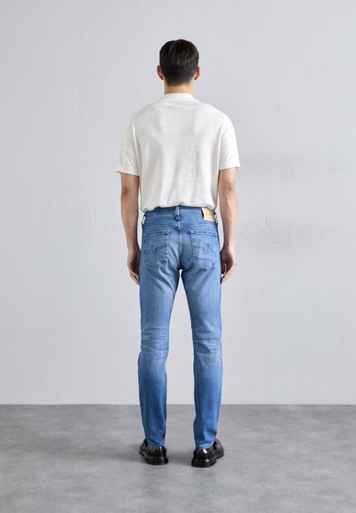 Svetlošedá košeľa s krátkym rukávom, modré džínsy s zadnými vreckami a vyblednutými detailmi, skombinované s čiernymi topánkami, zobrazené zozadu.