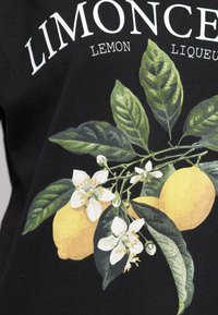 Svart t-shirt med ett livfullt grafiskt motiv av citron och vita blommor, med gröna blad, och fet vit text med orden "LIMONCÉ LEMON LIQUEUR."