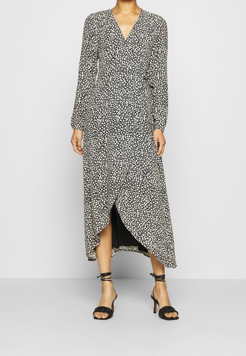 Femme portant une robe midi portefeuille à manches longues, noire et blanche à motifs floraux, avec un col en V et des sandales noires à talons hauts et à brides.
