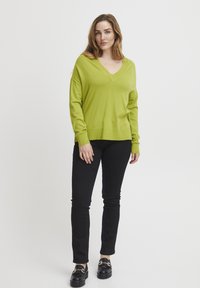 Limegrüner V-Ausschnitt-Pullover mit langen Ärmeln, kombiniert mit schwarzen Slim-Fit-Hosen und klobigen schwarzen Schuhen. Glatte Textur, lässiger Stil.