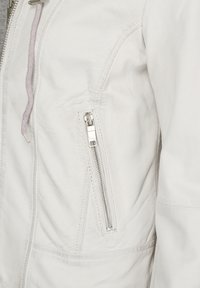 Weiße Lederjacke mit strukturierter Oberfläche. Verfügt über eine Reißverschlusstasche und einen kontrastierenden Kordelzug. Elegantes, tailliertes Design.