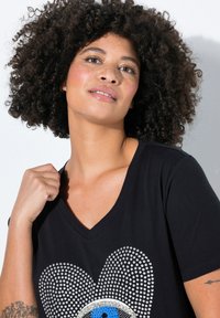 Schwarzes V-Ausschnitt T-Shirt mit einem Herzdesign aus silbernen Strasssteinen, das ein zentrales blaues Augendetail aufweist. Aus weichem Baumwollstoff, mit kurzen Ärmeln.