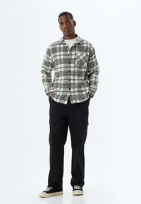 Camisa a cuadros en blanco y negro con dos bolsillos en el pecho, cierre de botones y mangas largas, combinada con pantalones cargo negros y zapatillas negras.
