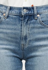 Κοντινή λήψη μπλε τζιν με κουμπωτό τζινάκι, θηλιές για ζώνη και χάλκινα καρφιά, φορεμένα με ένα λευκό πουκάμισο που φαίνεται μερικώς στην κορυφή.