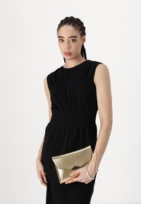 Zelta metāla maka ar aploksnes dizainu tur rokās. Kleita ir melna, bez piedurknēm, un tai ir savilkta detaļa vidukļa rajonā.