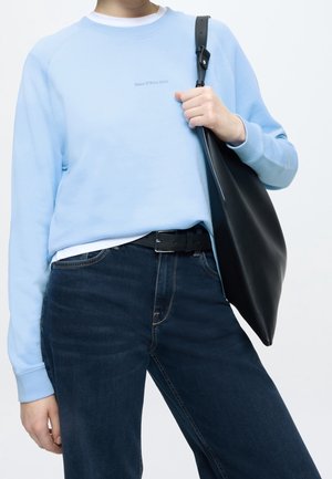 Personne portant un sweat-shirt bleu clair, un jean bleu foncé avec une ceinture noire, tenant un grand sac à épaule en cuir noir d'une main.