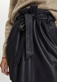 Svart läderkjol med en bred midjeband och en stor knuten rosett. Tyget har en jämn textur och är skräddarsytt med veck.