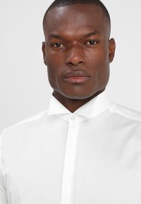 Weißes Hemd aus glattem Stoff mit Knopfleiste und klassischem Kragen. Der Stoff wirkt leicht und gut sitzend.